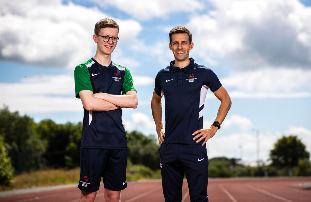 Oliver Gunning and guide Kyle Duncan are standing on a running track. Oliver stands on the left and has his arms crossed. Kyle has his hands on his hips. They are wearing their Commonwealth Games training kit.